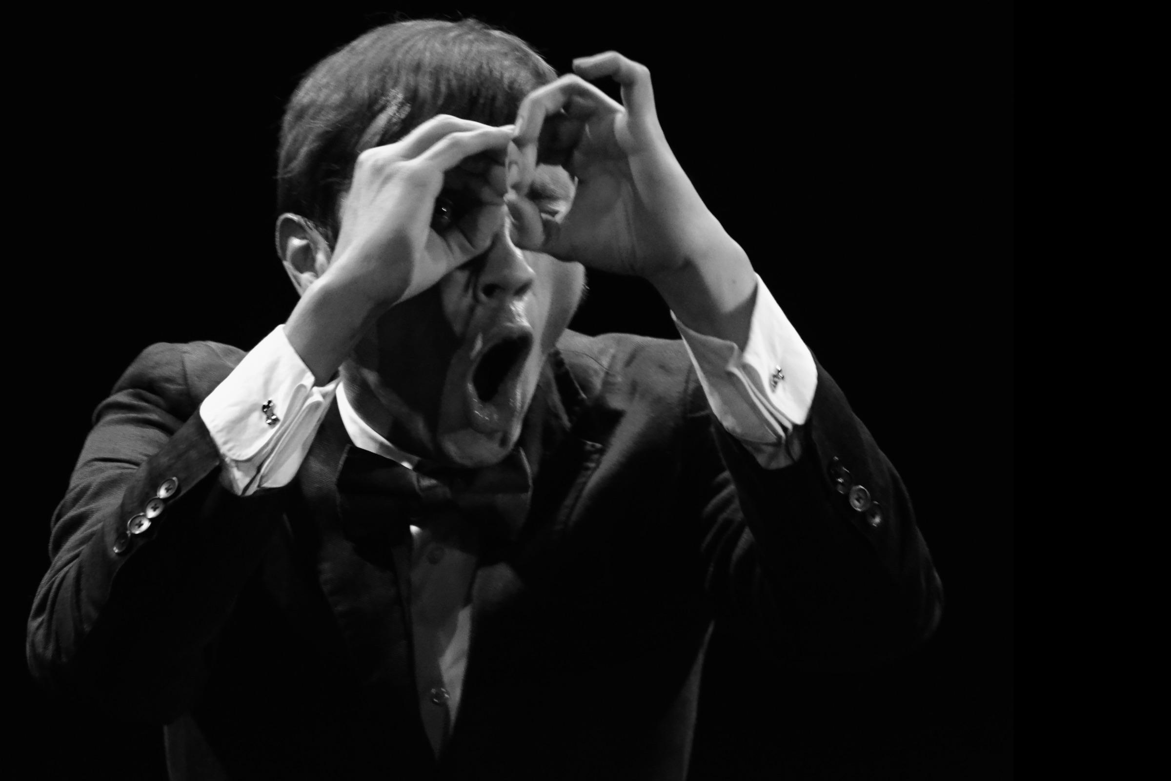 Black-and-white photo of Caspar Schjelbred performing physical improvisation on stage, forming expressive binoculars with his hands and reacting with a wide-open mouth.